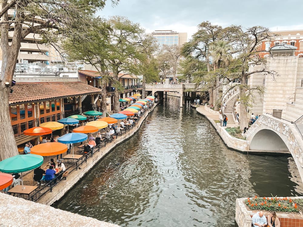 A view of the San Antonio River Walk showcasing multicolored umbrellas, a serene waterway, and a bridge built from brick and stone with trees lining the waterway.