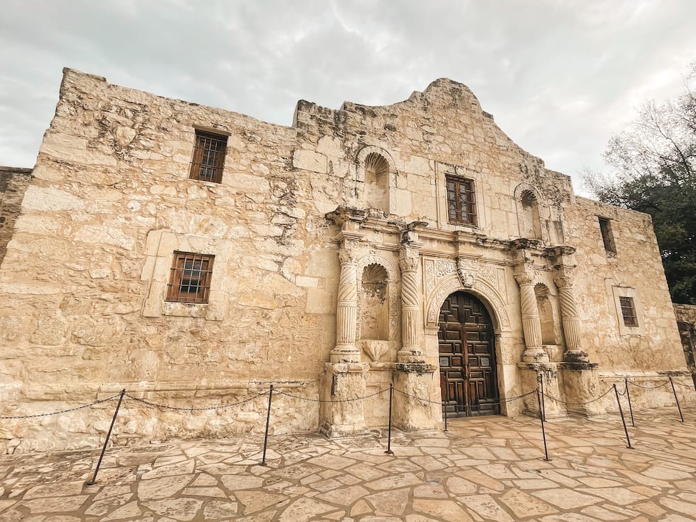 A close-up view of the outside of The Alamo, a Spanish mission from the 1800s that features old-looking tan stonework, a wooden door, and iron windows.
