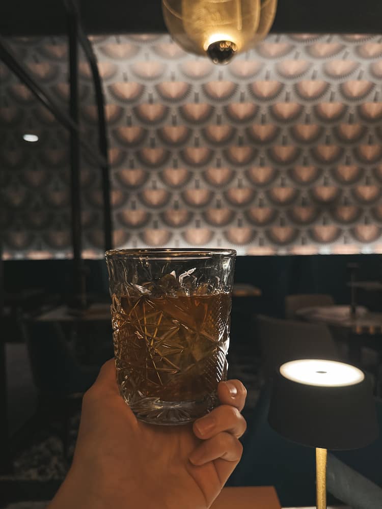 A bourbon cocktail in a textured glass in front of a pink and grey wallpapered wall in a dark speakeasy in Scottsdale.