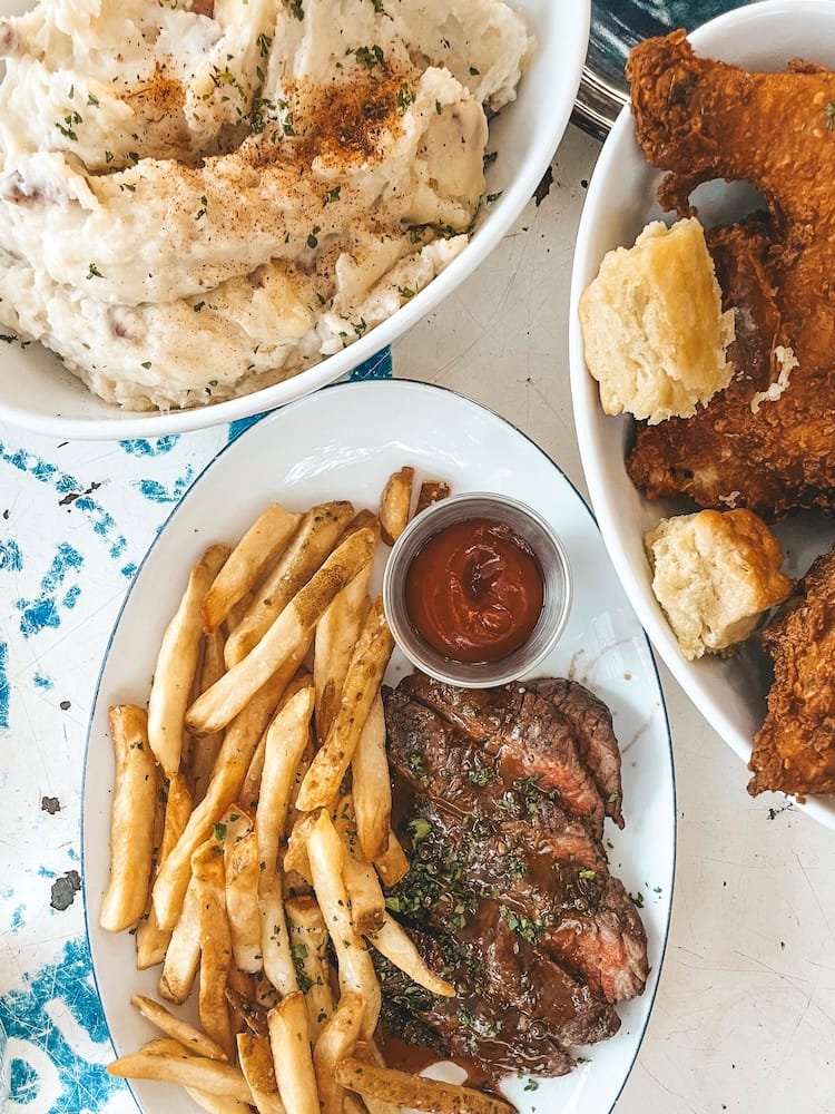 Several plates of food sitting on a table, including steak frites and French fries, fried chicken, and mashed potatoes.