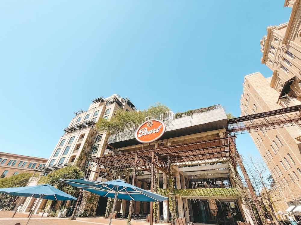 A historic industrial-style building with a red sign that says "Pearl" in white cursive letters and blue umbrellas in the front