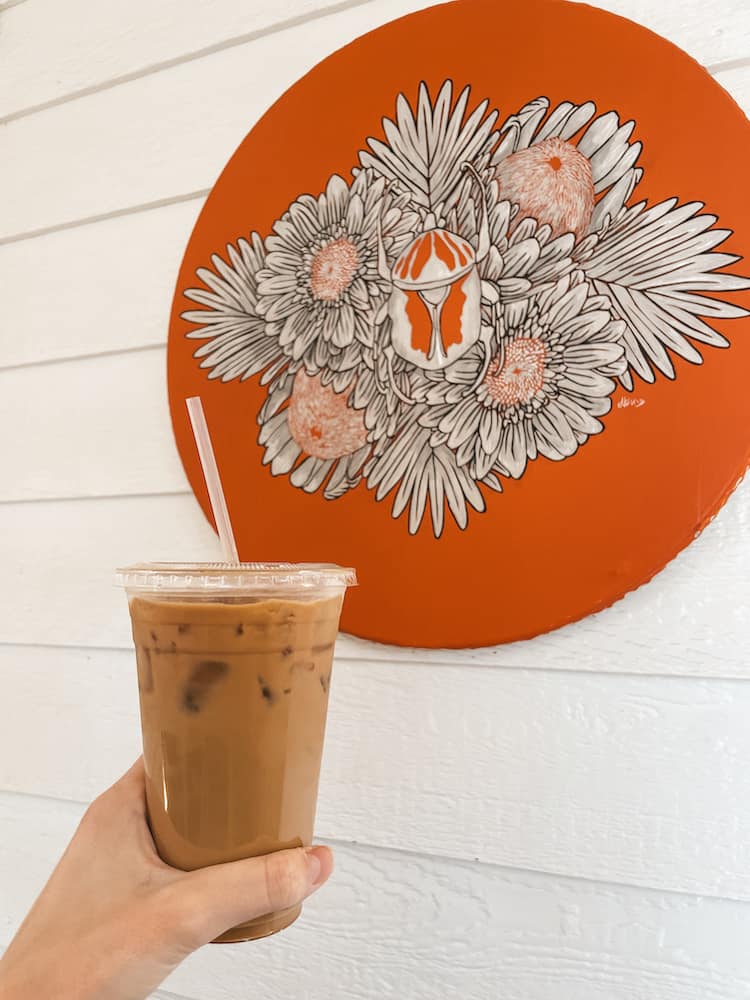 An iced cup of coffee in front of a red floral sign on a building with white siding.
