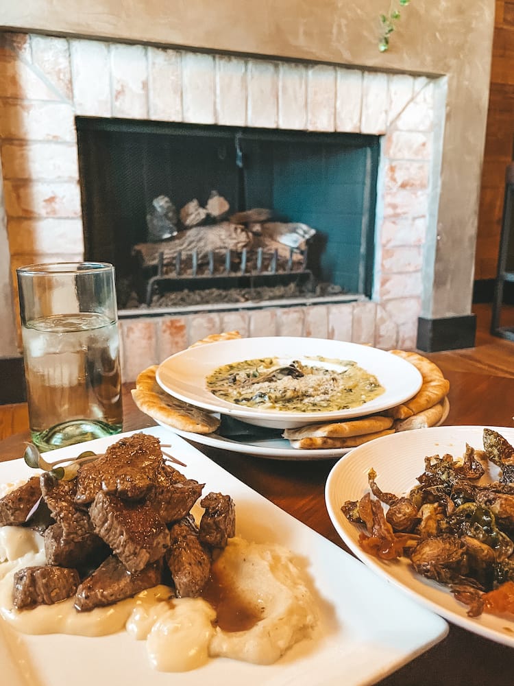 Several plates of food, including steak tips, Brussels sprouts, and spinach artichoke dip, in front of a fireplace