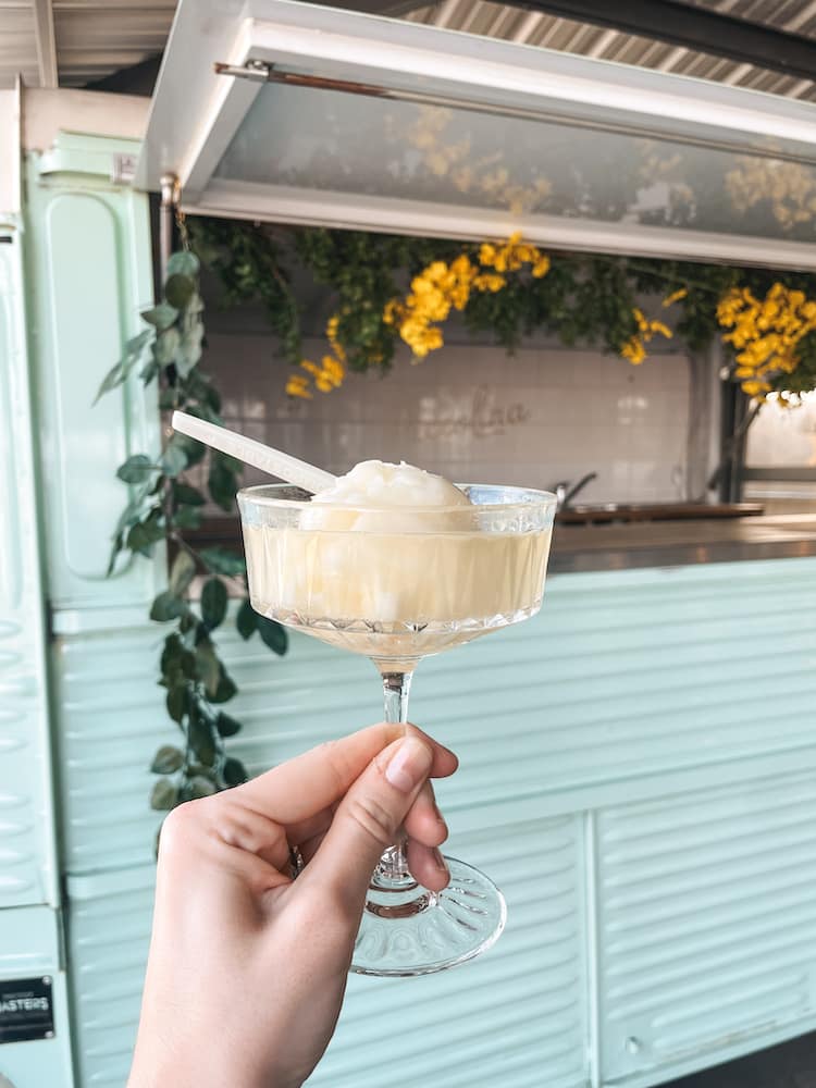 A scoop of Italian shave ice in a coup glass in front of a mint green cart with yellow flowers and greenery in it.