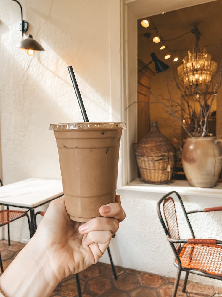 An iced mocha being held up inside a coffee shop with white stucco walls and potted plants.