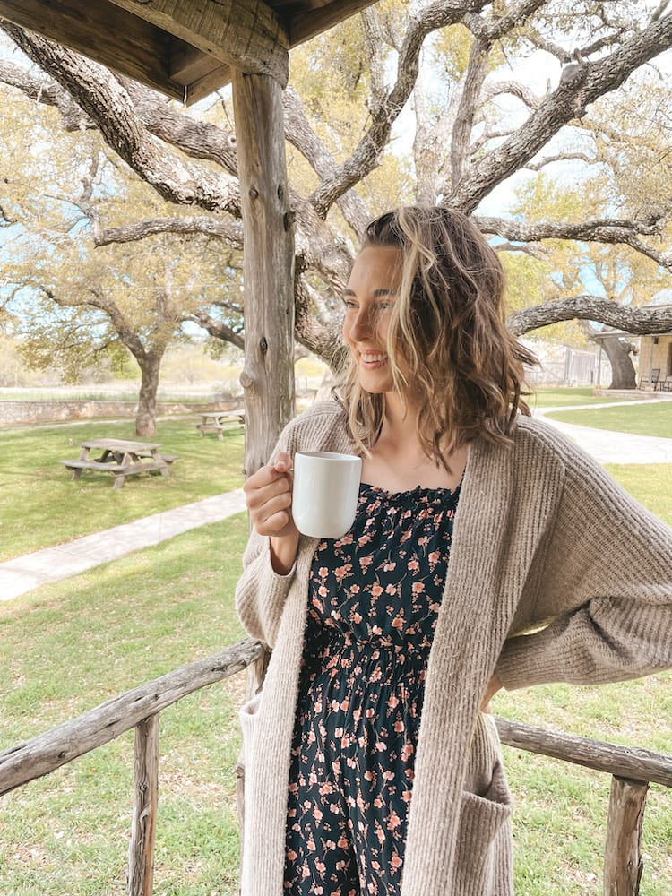 A woman wearing a black jumpsuit and sweater holding a white mug of coffee and looking into the distance on a wooden porch with trees and grass in the background.