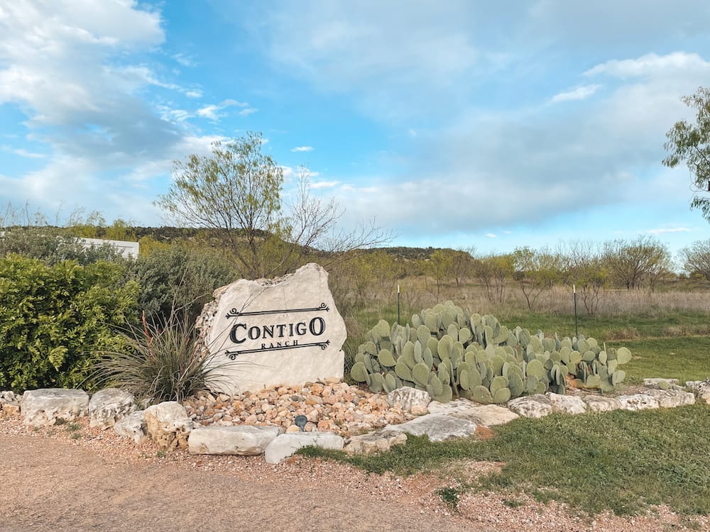 The sign to Contigo Ranch on a granite rock surrounded by cactus, trees, and other desert plants.