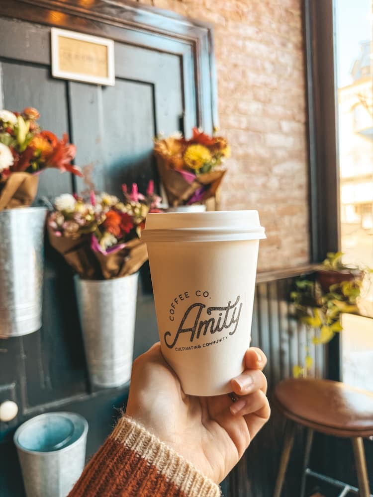 A cup of coffee from Amity Coffee Co in Penn Yan being held in front of a brick wall with several metal buckets holding colorful flowers.
