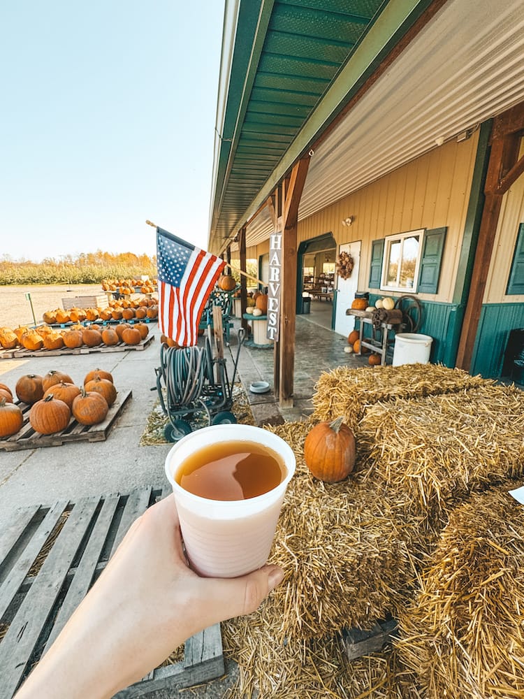 A cup of apple cider is being held in front of a farm stand with hay bales, pumpkins, and an American flag out front in Upstate New York in October.