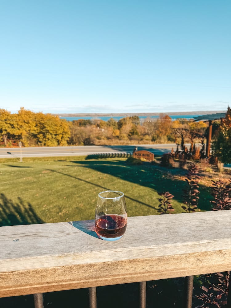 A glass of red wine sitting on a wooden ledge overlooking a grassy field with fall foliage and a blue lake in the background