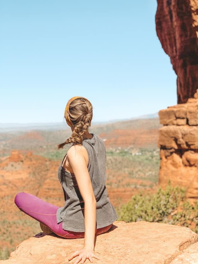 A woman wearing a gray tank top, pink leggings, with braided hair sitting on the ledge of one of the red rocks in Sedona on Cathedral Rock Trail