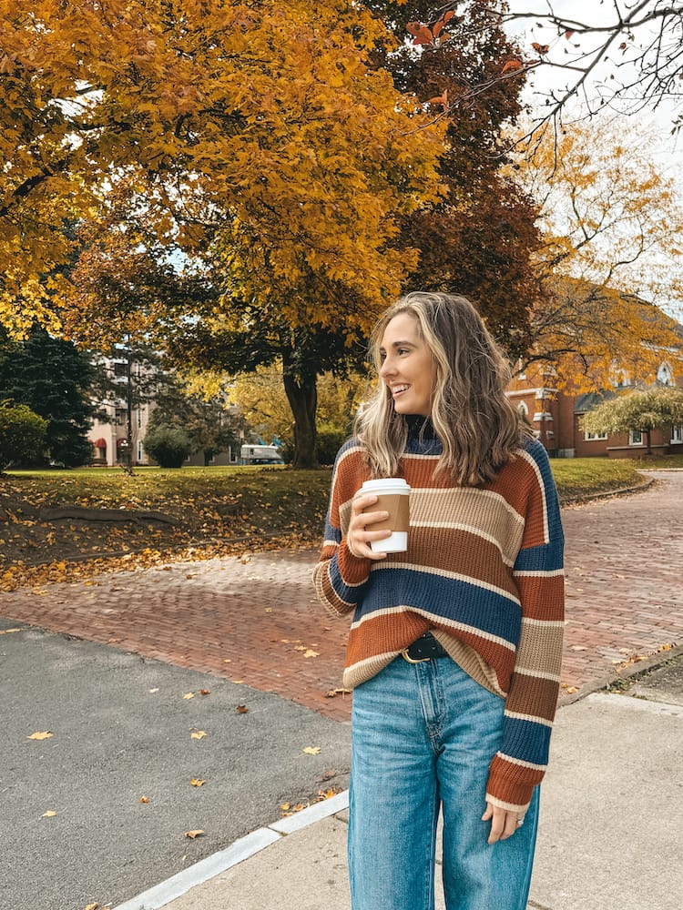 A woman in a striped sweater and jeans standing in front of a fall tree in Geneva, one of the best towns in the Finger Lakes.