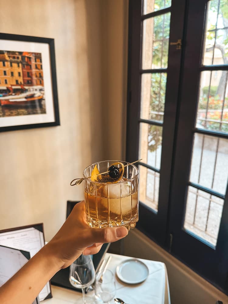 An old fashioned in a cocktail glass being held in front of a window and artwork hanging on the wall at a fine dining restaurant in Sedona.