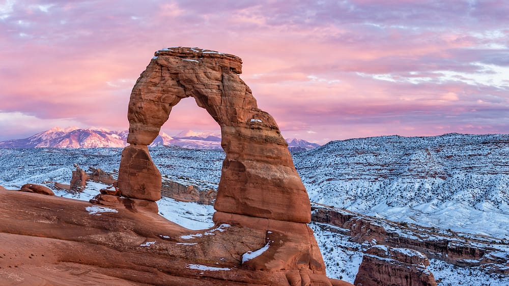 Delicate Arch in Arches National Park with a bright pink sunrise in the background and snow-dusted red rocks surrounding it.