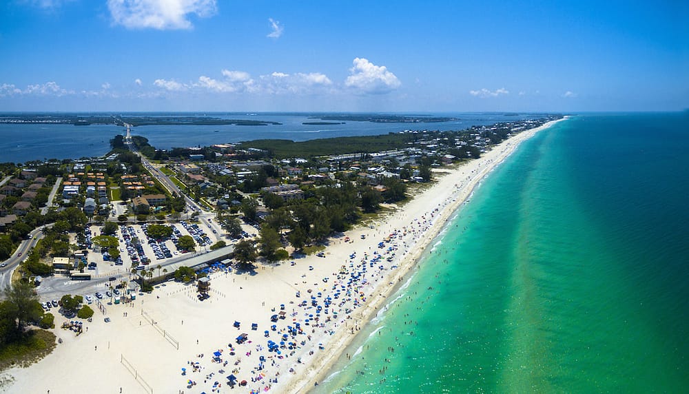 An aerial view of the bright blue water in Anna Maria Island and long stretch of white sand beaches dotted with blue umbrellas.