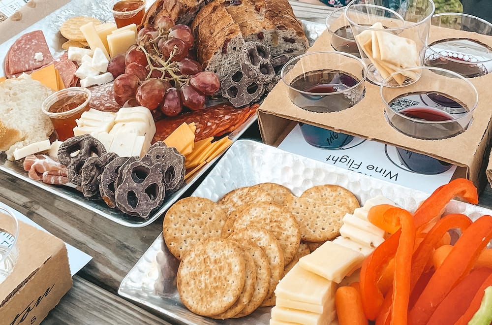 A flight of four wines and grapes, pretzels, cold cuts, veggies, and other snacks on a table