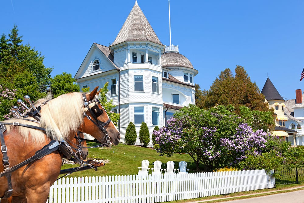 A horse-drawn carriage pulling up in front of a white Victorian house with a white picket fence in Mackinac Island, MI, which is one of the best places to visit in the USA in 2023.