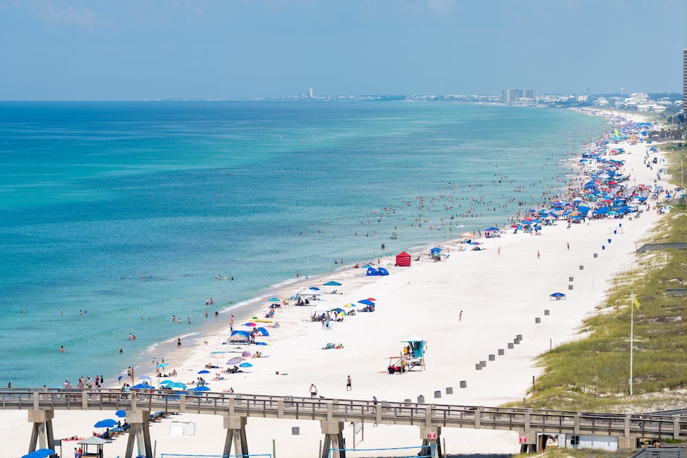 A busy day at Panama City Beach with several blue, red, and multicolored umbrellas pitched in the white sand in front of the turquoise waves.