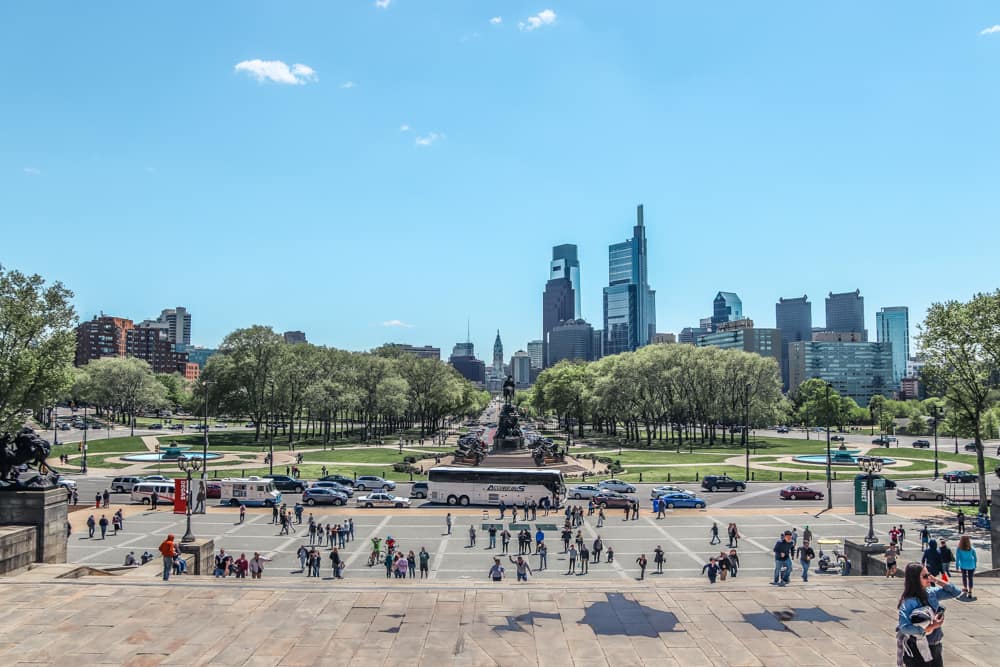 A view of the Philadelphia skyline with cars, tour buses, and people roaming about with a clear blue sky.