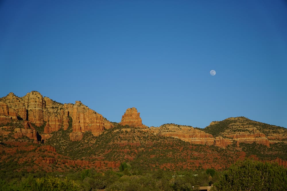 A view of the red rocks in Sedona with an outline of the moon in the sky at dusk.