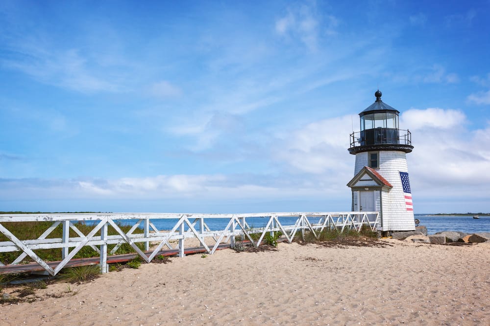 A short white lighthouse with an American flag painted on the side on a beach in Nantucket.