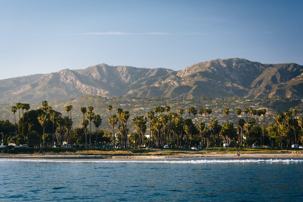 A view of the coast in Santa Barbara, with the ocean and palm trees in the foreground and the mountain range in the background.