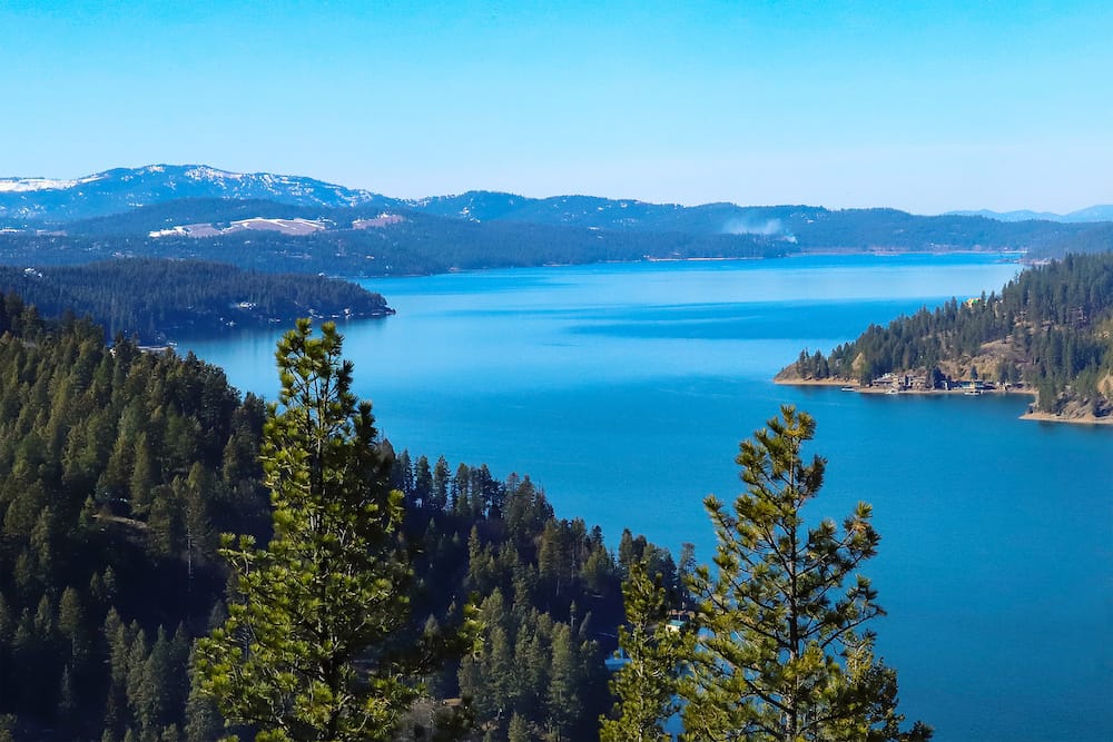 A blue lake surrounding by green pine trees in Coeur D’Alene, one of the best places to visit in the USA in August.