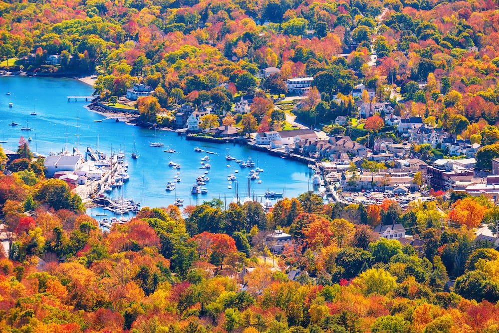 The vibrant fall foliage surrounds the blue water and boats in the harbor in Camden, Maine.