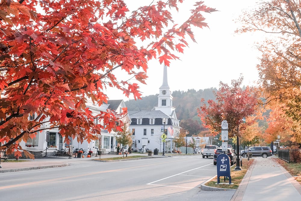 A white church in Stowe, Vermont – one of the best places to visit in the USA in October – surrounded by red, orange, and yellow fall leaves and trees.