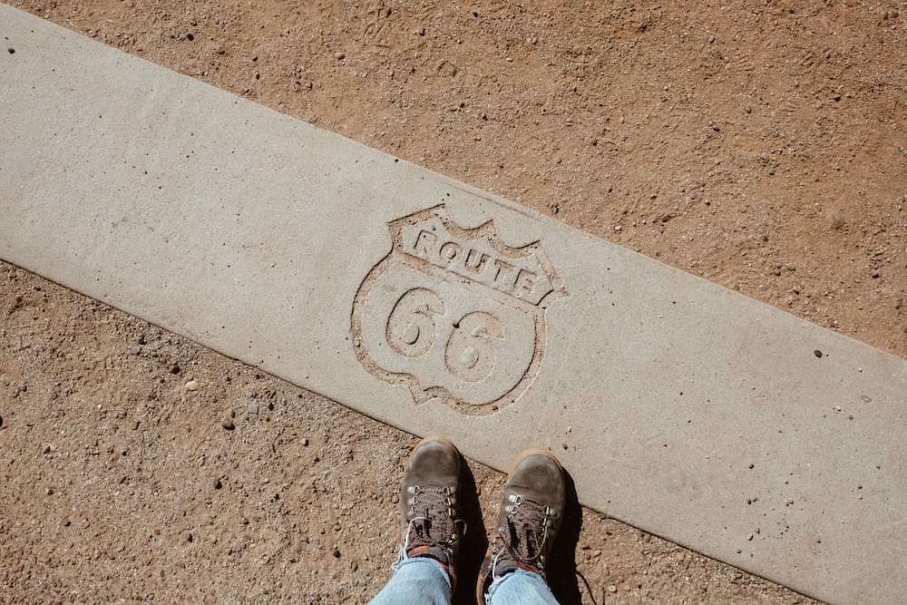 A pair of black boots standing by the Route 66 sign on a dirt path.