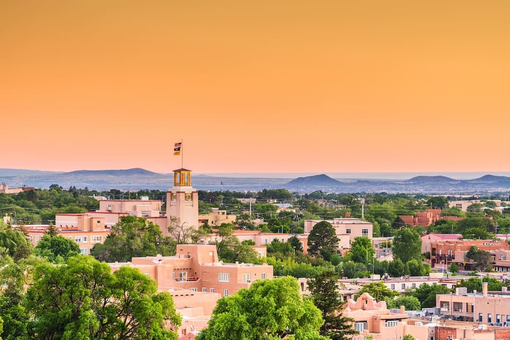 The skyline of Santa Fe, New Mexico, during the sunset, with adobe-style red buildings dotted throughout the town and an orange sky.