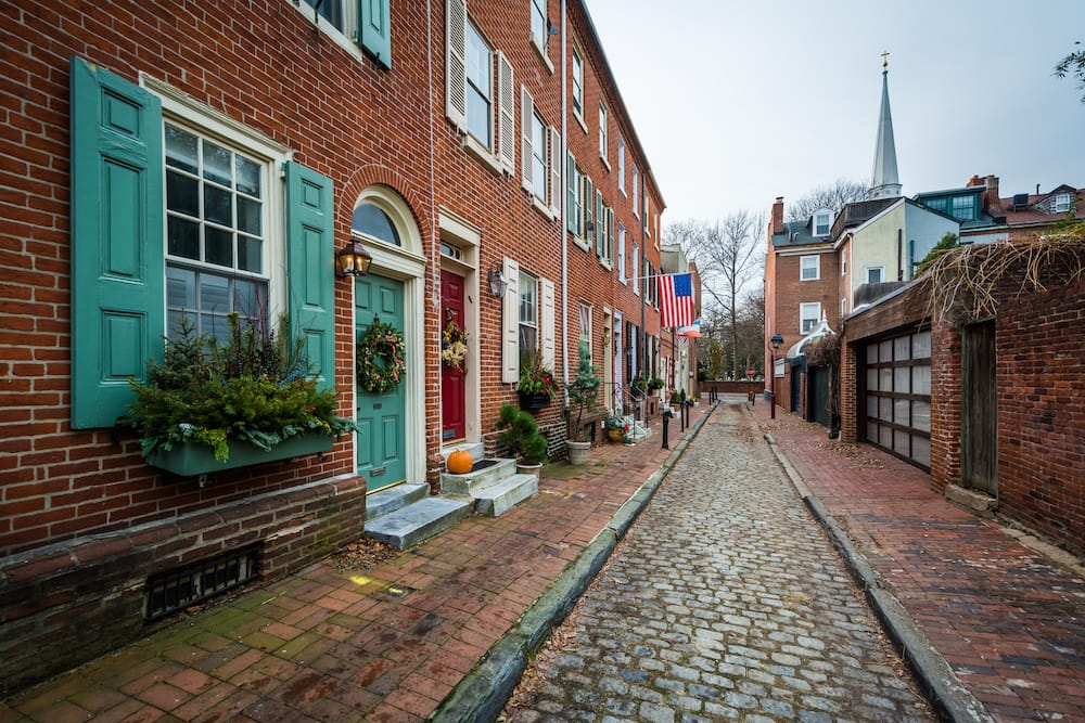 A red brick and cobblestone alleyway in Philadelphia with green and red doors adorned with wreaths and greenery for the holidays.
