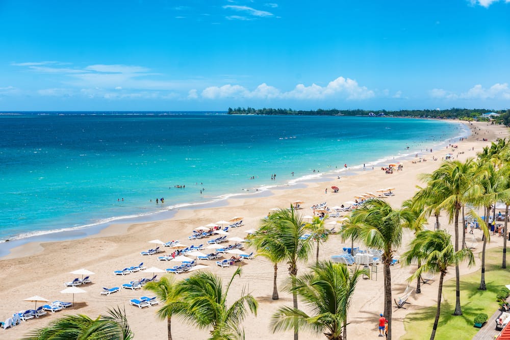 Green palm trees, a golden sand beach, blue beach chairs, and several people overlooking the clear, blue ocean water in San Juan, Puerto Rico.