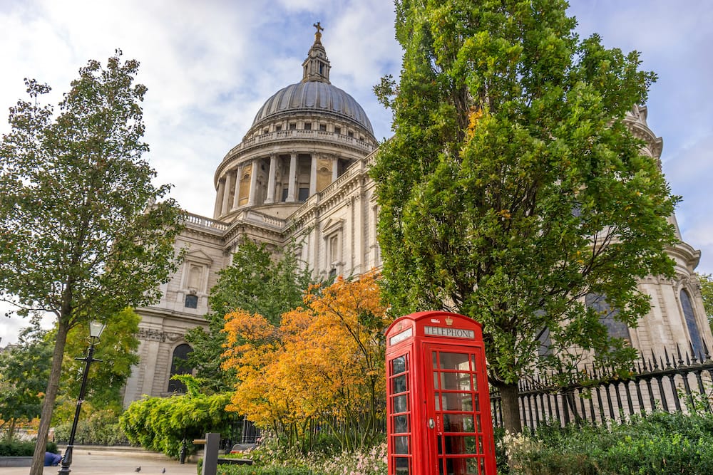 A red telephone booth in front of changing green and yellow trees during the fall with St. Paul's Cathedral in the background.