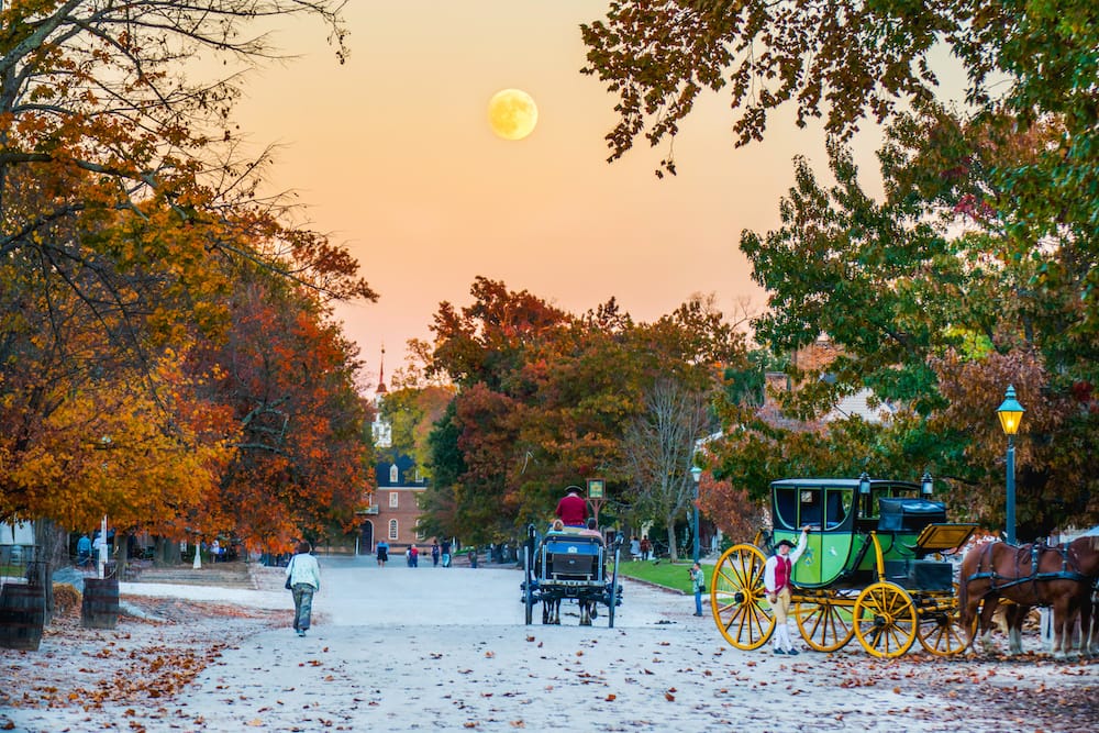 Two horse-drawn carriages driving through the snowy streets of Colonial Williamsburg at the end of the fall, with the leaves changing colors in the background.