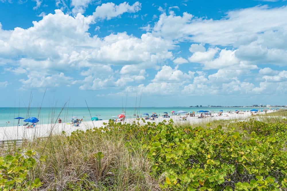 An overcast day at Pass-a-Grille Beach, with dozens of fluffy while clouds in the blue sky. Greenish-blue water laps against the white sand beach, where dozens of beachgoers with with colorful umbrellas. Green plants and grasses sit in the foreground.