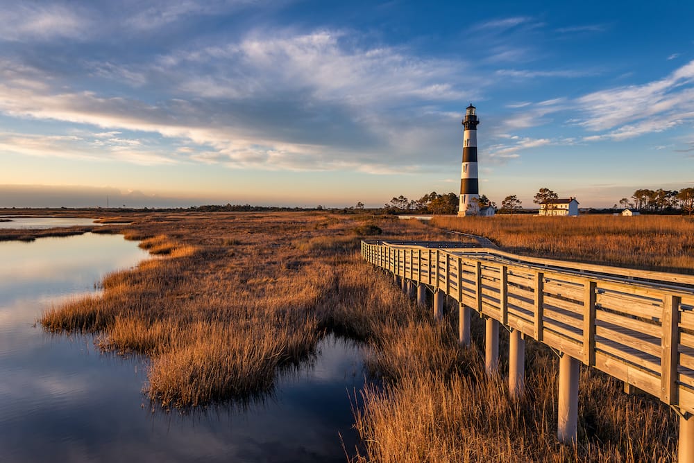 A black and white striped lighthouse at the end of the pier in a marshy swamp in Outer Banks.
