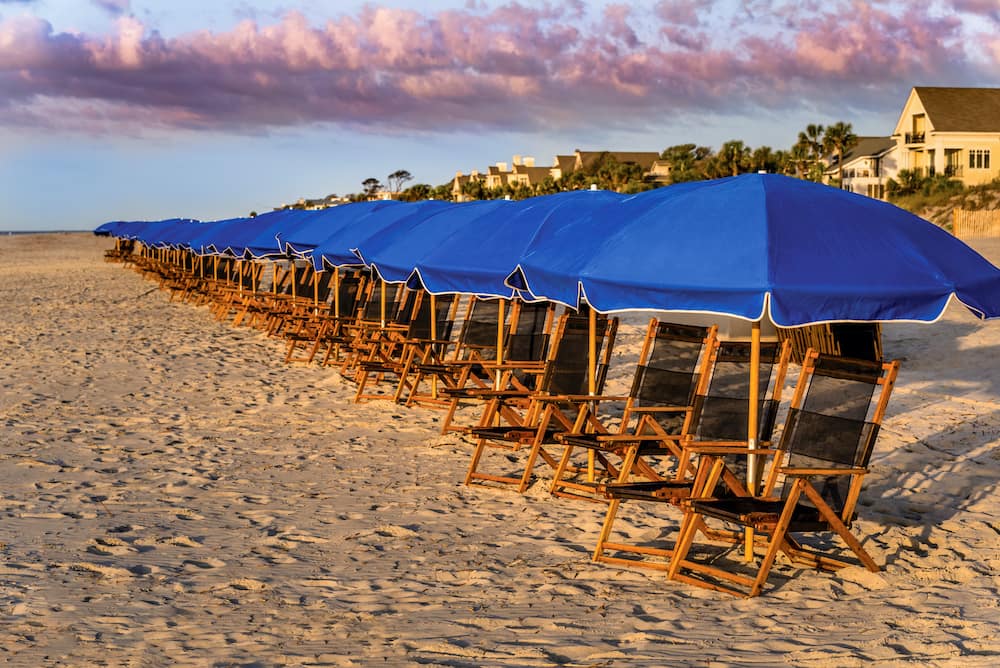 A link of beach chairs with blue umbrellas sitting on the sand at sunset on Hilton Head Island - one of the best summer beach vacations in the U.S.