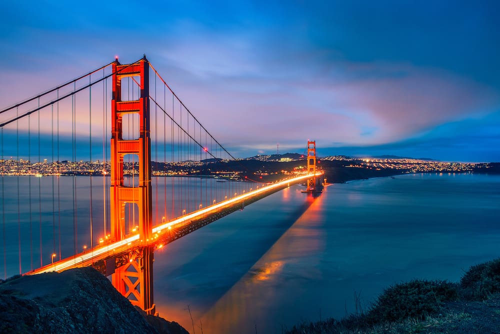 The red Golden Gate Bridge spanning across a body of water at dusk, with city lights glimmering in the background and a tinge of the pink sunset in the night sky.