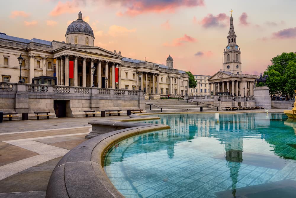 The National Gallery and other buildings in Trafalgar Square reflecting in a pool of blue water at sunset.