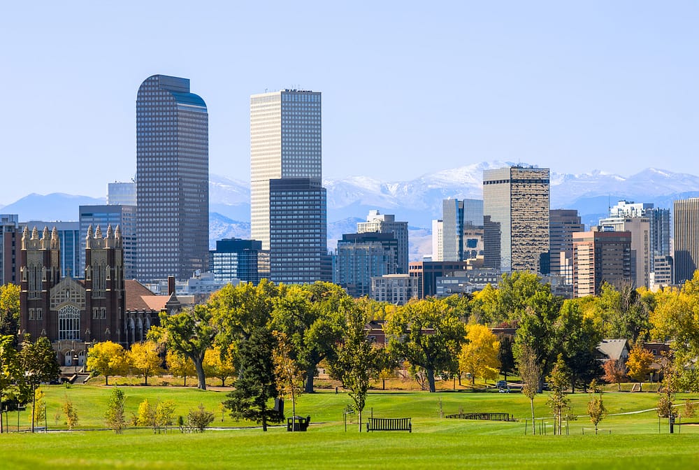 The skyline in Denver, Colorado, with green trees and a grassy park in the foreground.