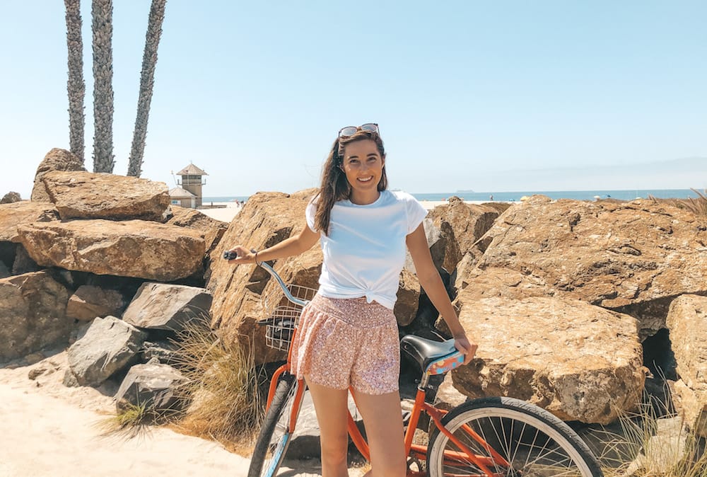 A girl holding a bike behind her is standing in front of rocks and the beach. Best Things to Do on Coronado Island | Travel by Brit
