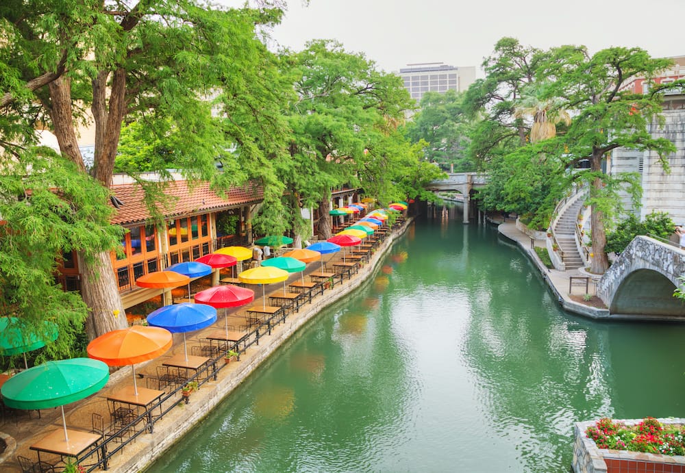 Several colorful umbrellas and green trees next to the blue river of the San Antonio Riverwalk.