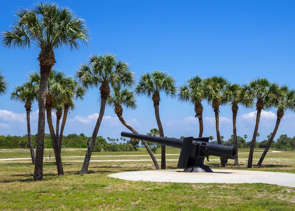 A black cannon sitting in the middle of Fort De Soto Park – one of the best cheap things to do in St. Pete – surrounded by green palm trees, grass, and a blue sky