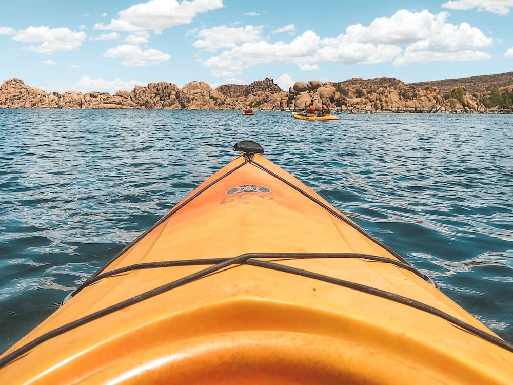 An orange kayak floating toward reddish rock formations in a blue lake in Prescott, AZ, one of the best places to visit in the USA in April.
