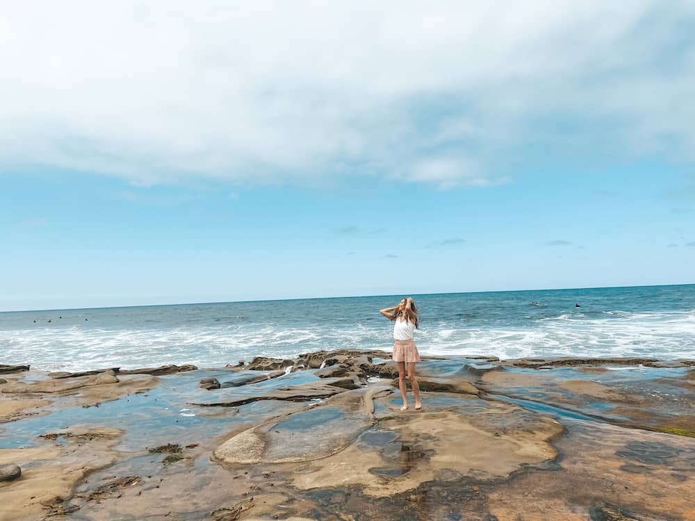 A girl standing on rocks with the blue ocean and blue sky in the background.