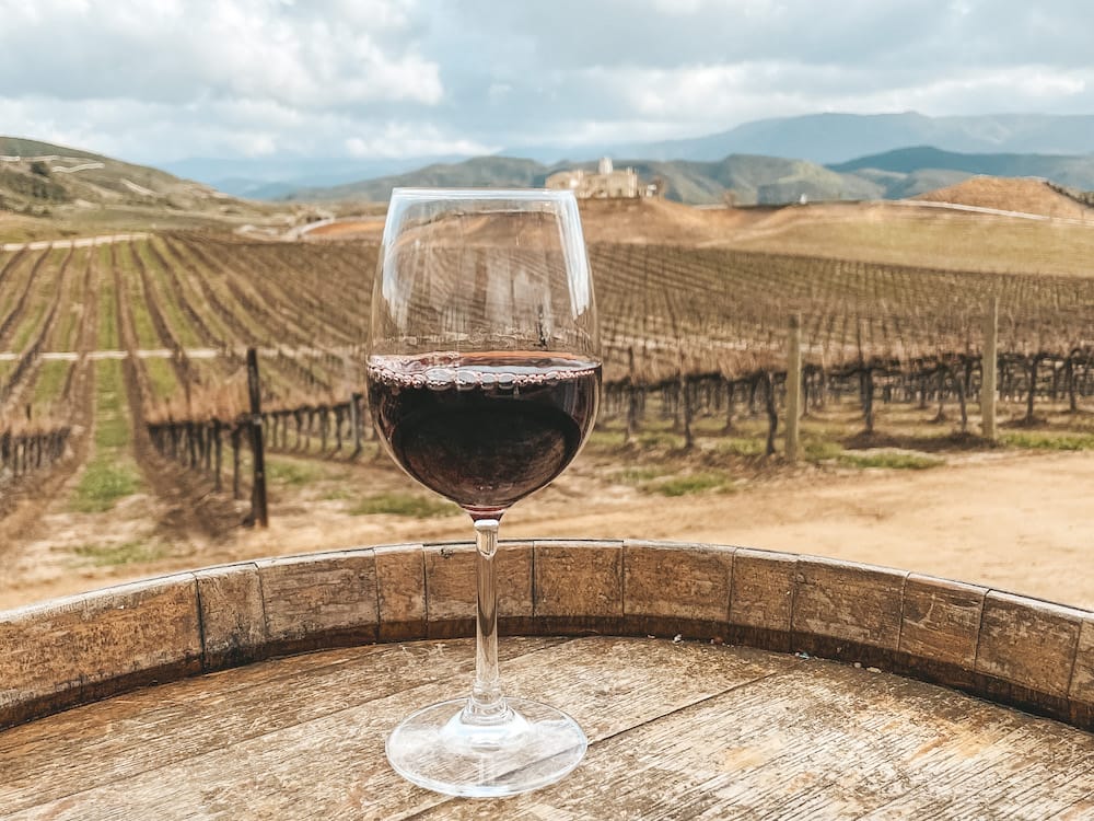 A glass of red wine sitting on a wooden barrel in front of a dead vineyard during a wine tour in Temecula.