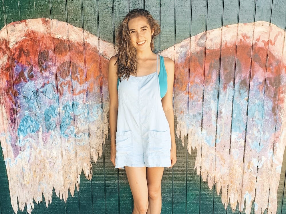 A girl with brown hair wearing a green swimsuit and denim overalls standing in front of pink, blue, and purple wings painted on the side of a green building in Haleiwa Hawaii.