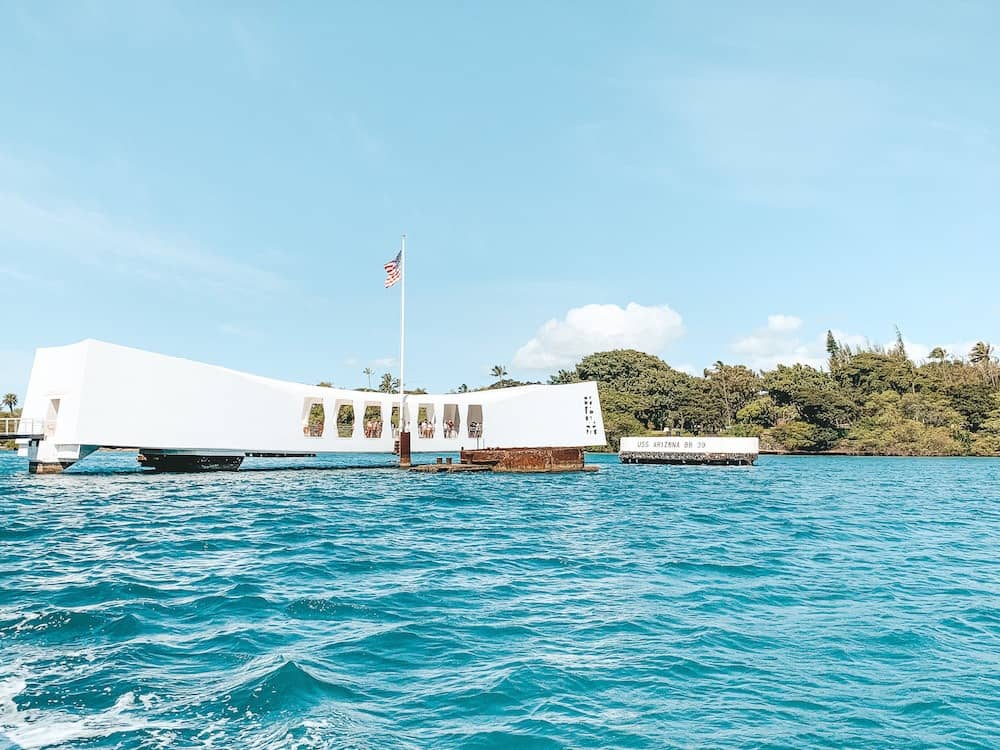 The USS Arizona Memorial at Pearl Harbor National Monument in Honolulu on the island of Oahu. The memorial is a long, white building floating on the blue island water. This is an American flag flying from the center of the memorial.