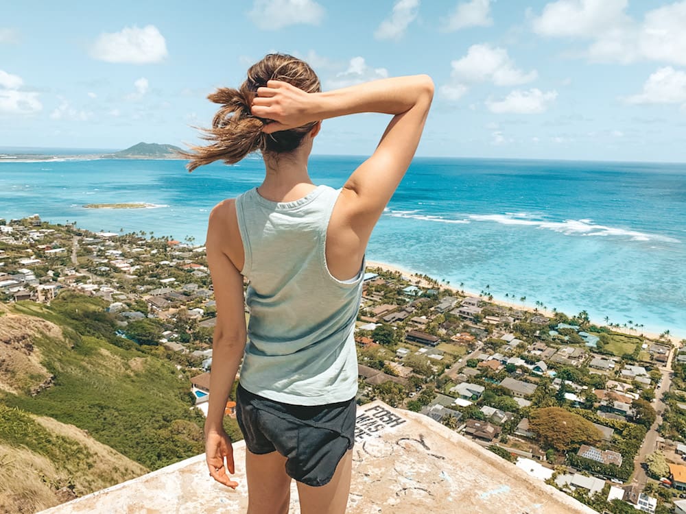 A girl with a brown ponytail wearing a green tank top and black athletic shorts standing on the edge of a pillbox overlooking the Pacific Ocean and a neighborhood on the island of Oahu at the end of the Lanikai Pillbox Trail in Hawaii.